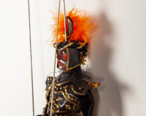 Marionette puppet of a man with a mustache wearing gold and black clothes with orange feathered hat.