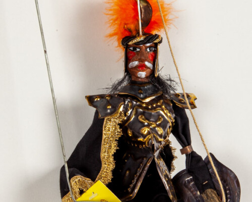Marionette puppet of a man with a mustache wearing gold and black clothes with orange feathered hat.